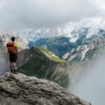 man with red backpack standing on cliff facing mountains under white sky during daytime